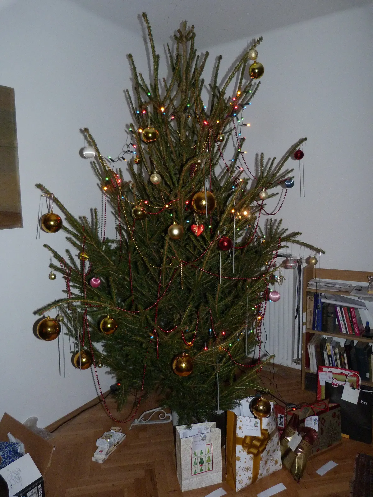 Real Christmas tree with gold baubles and gifts in a living room corner next to a bookshelf