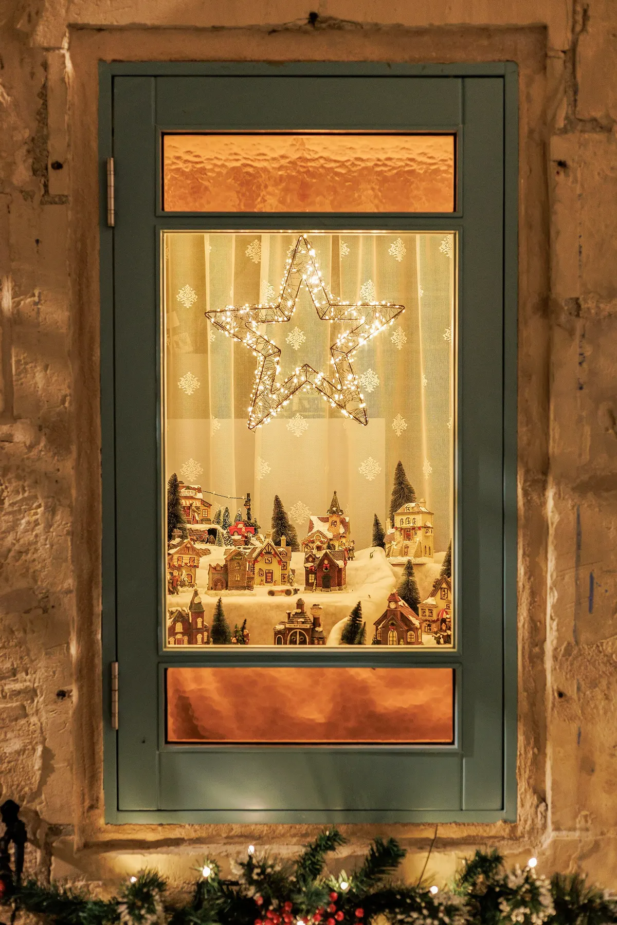 Window glowing with a fairy-light star above a miniature snowy village and a sheer curtain