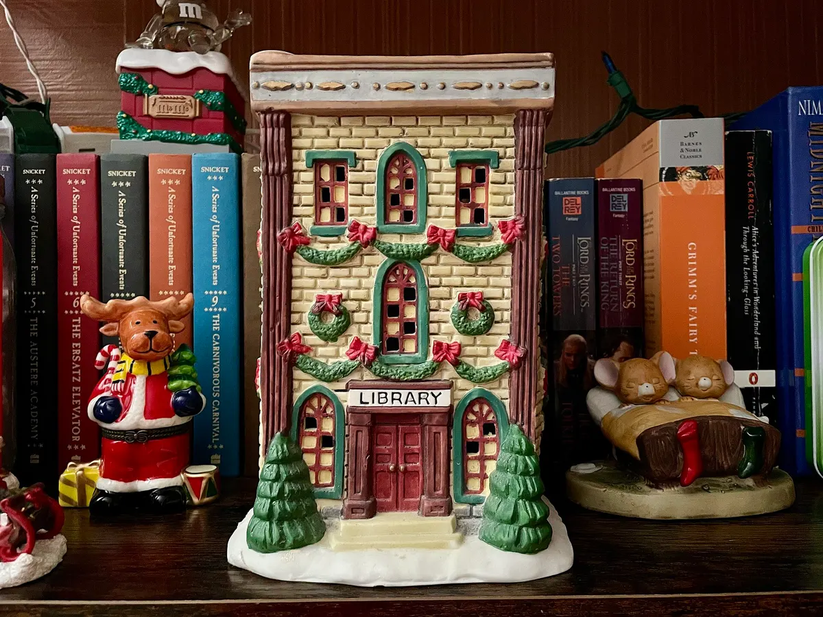 Ceramic Christmas village library building and small figurines on a bookshelf with green string lights