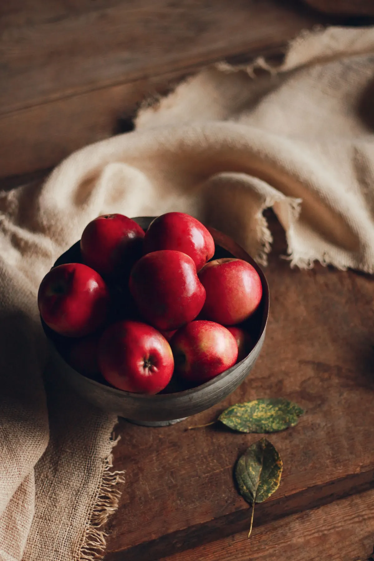 Dark gray metal bowl of red apples on a wooden table with frayed burlap and two leaves beside it
