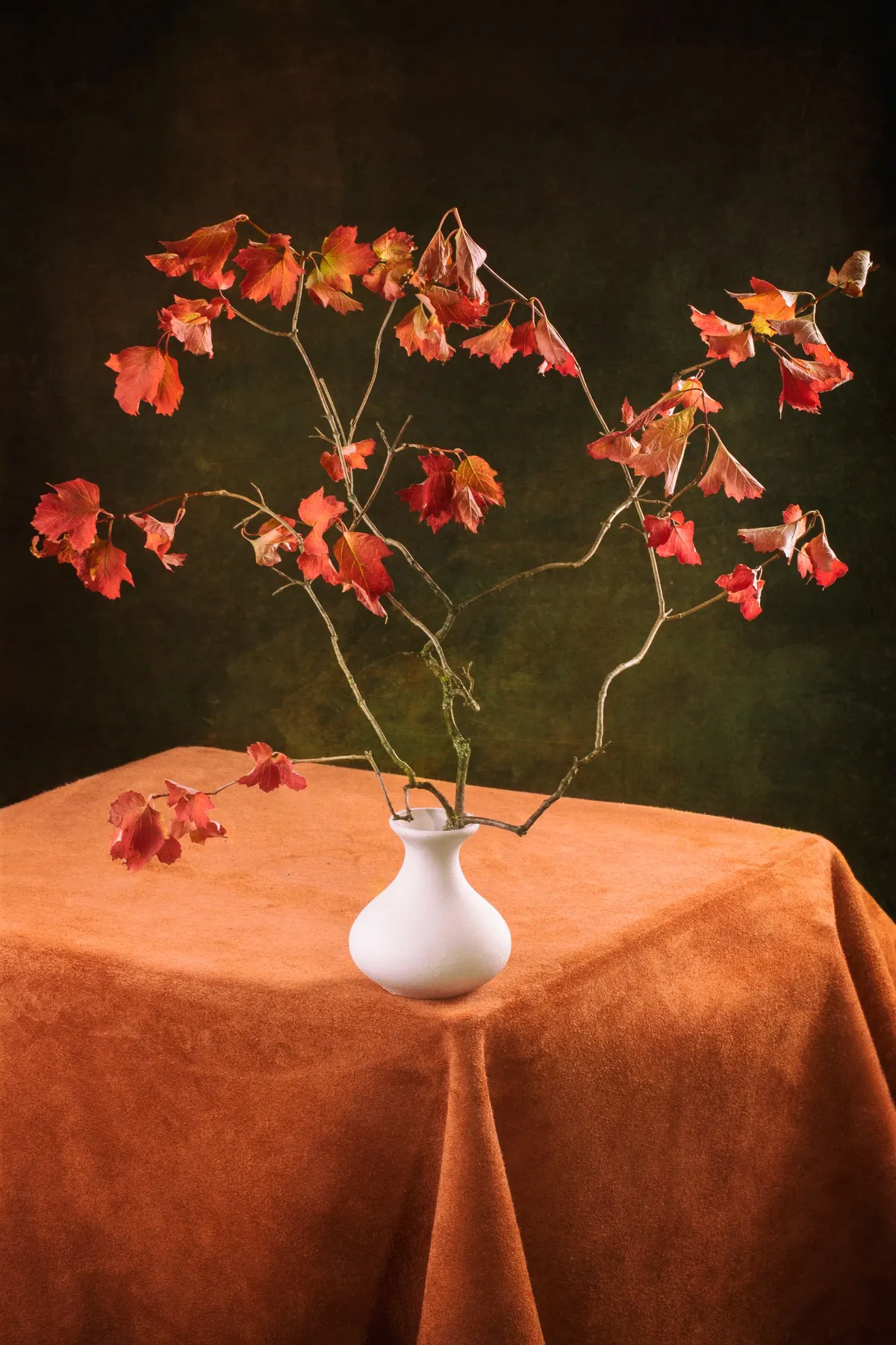 White vase of thin branches with red, orange and brown leaves on a table covered in rust-colored fabric