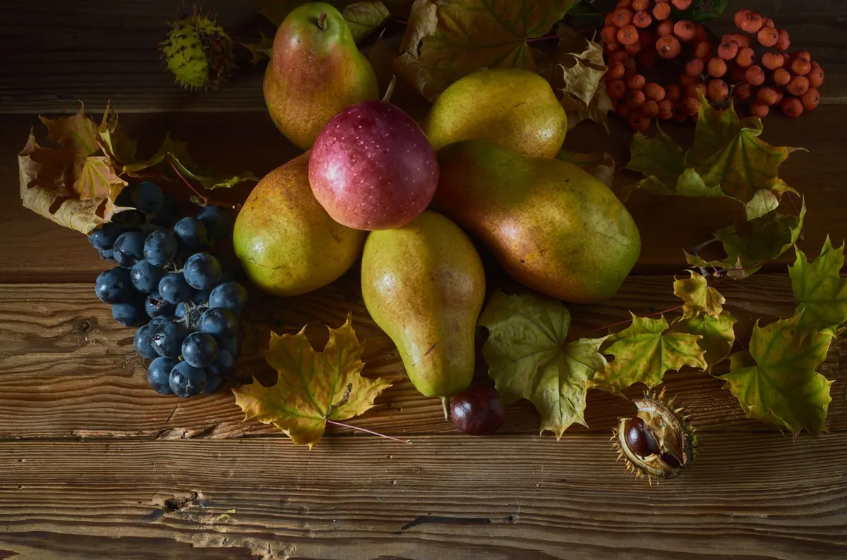 Yellow-green pears, a red apple, dark grapes, orange berries, chestnuts and autumn leaves spread across dark wooden planks