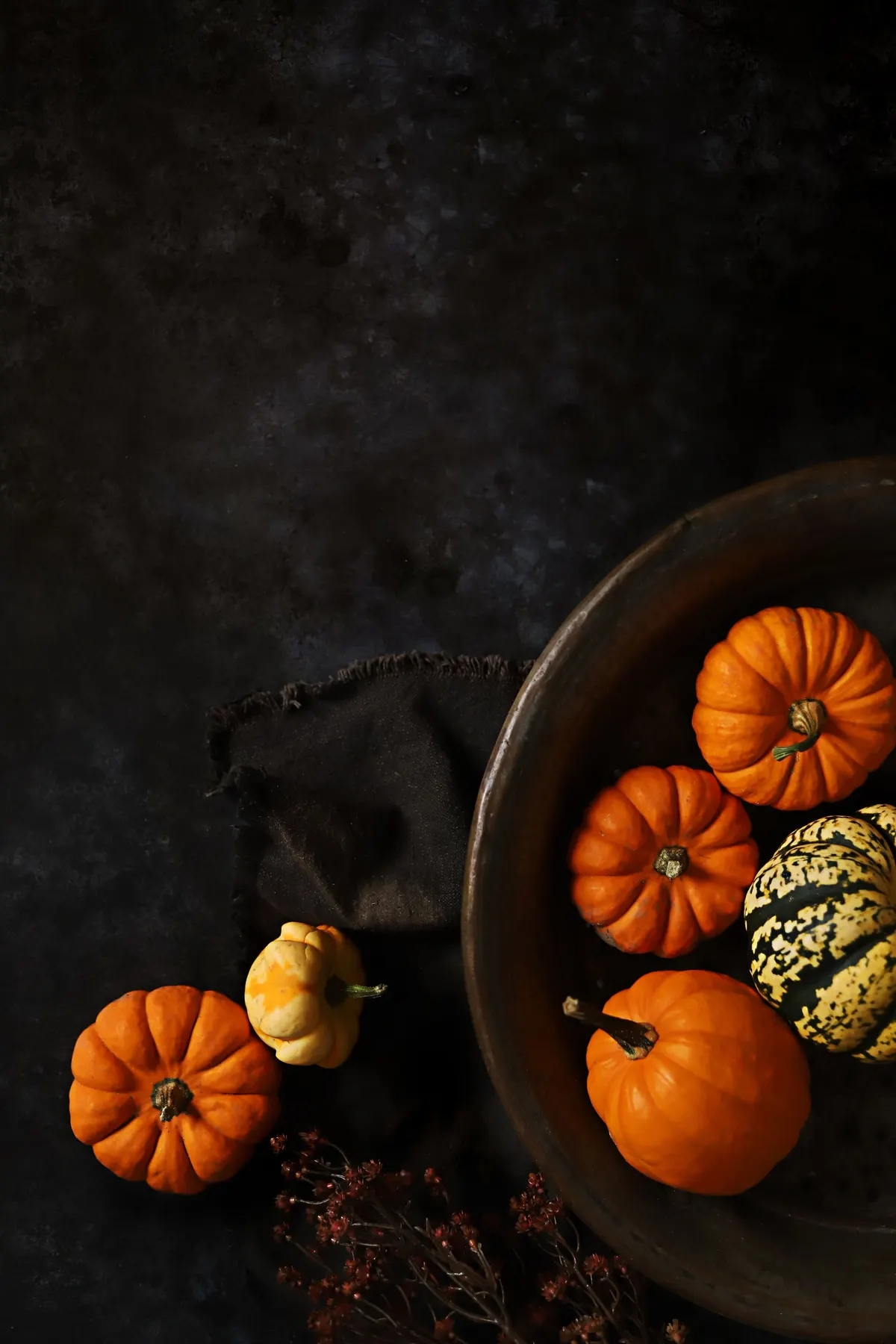 Large brown bowl holding orange pumpkins and a cream and green striped gourd on a dark fringed cloth