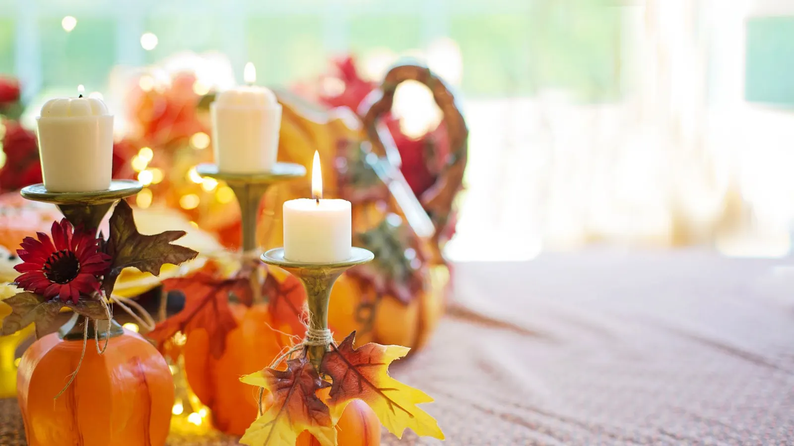 Four orange pumpkins with three cream candles in brass holders, scattered autumn leaves and string lights on a patterned table runner
