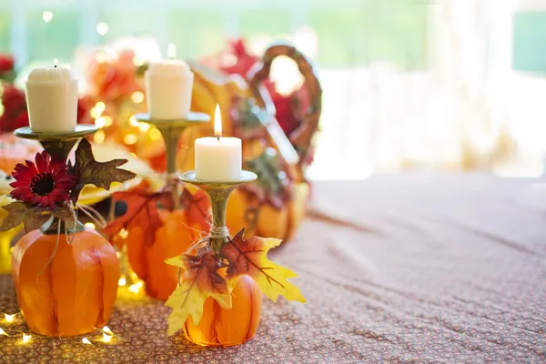 Four orange pumpkins with three cream candles in brass holders, scattered autumn leaves and string lights on a patterned table runner