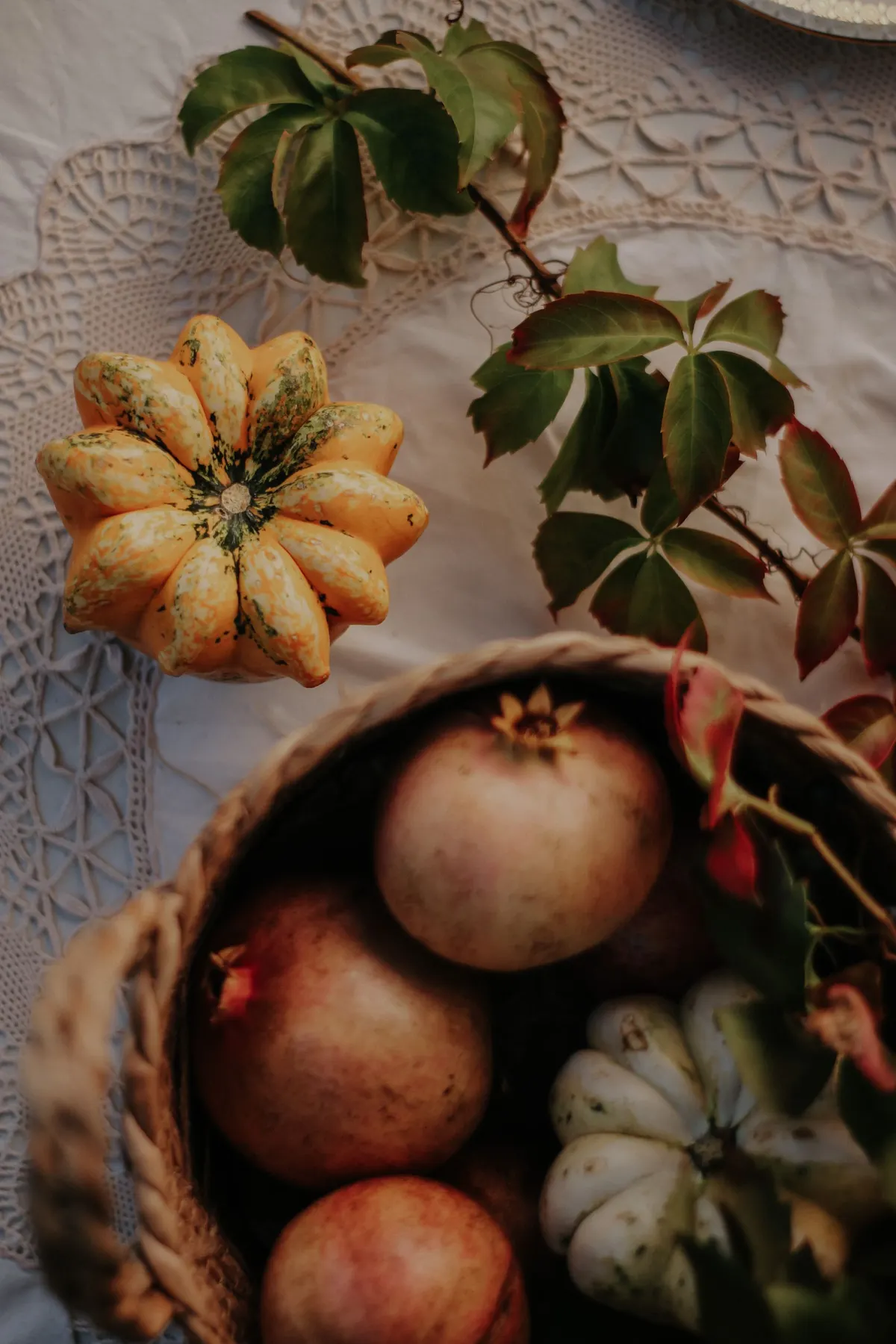 Two ribbed gourds, three pomegranates and a wicker basket with leafy vines on an ivory lace tablecloth