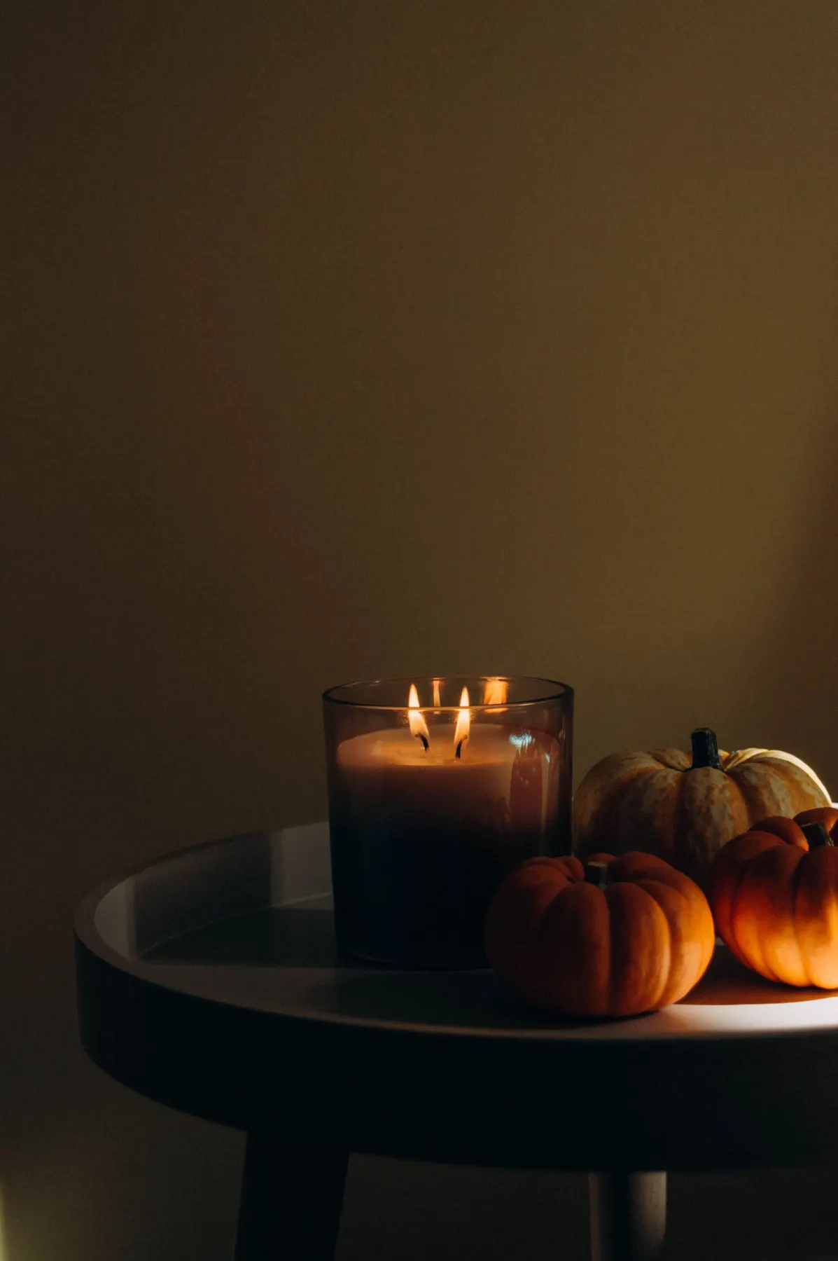 Three small pumpkins and a lit two-wick candle in a dark glass jar on a small round dark table