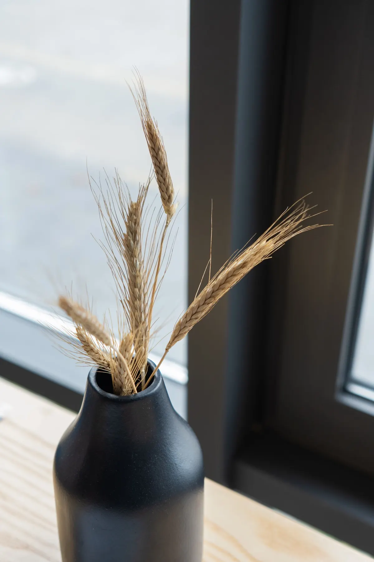 Matte black vase holding golden dried wheat stems on a pale wooden surface beside a dark-framed window