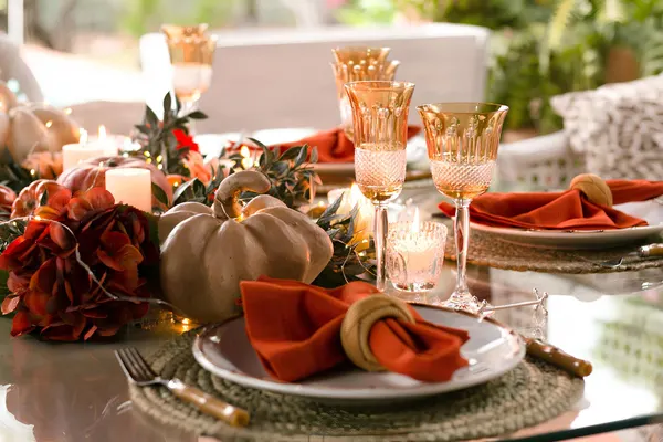 Glass-top table with a low gray-green pumpkin and red flower centerpiece, white pillar candles, amber goblets and rust napkins