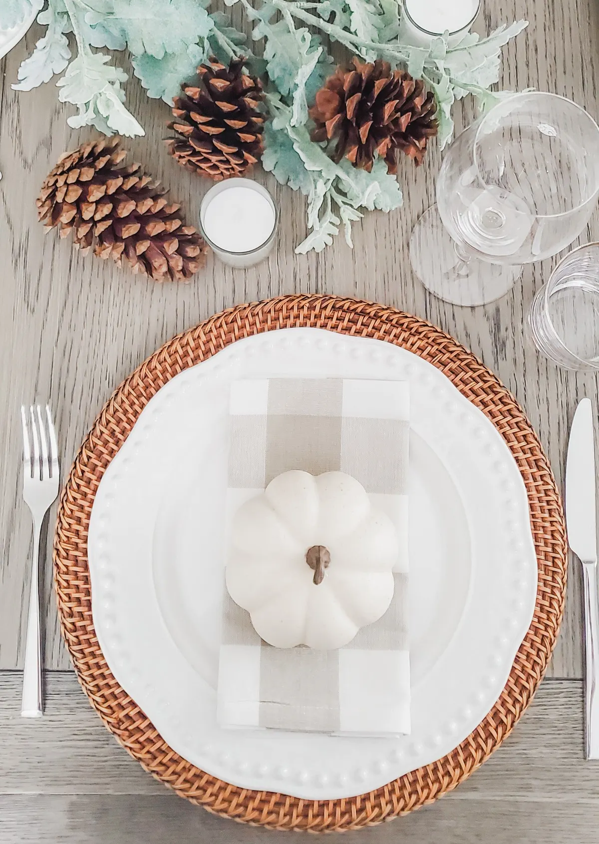 Overhead place setting with a white plate on a rattan charger, buffalo-check napkin, white mini pumpkin, pinecones and sage sprigs
