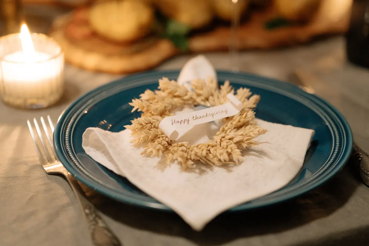 Dark teal plate holding a folded white napkin, a small dried wheat wreath and a handwritten scroll