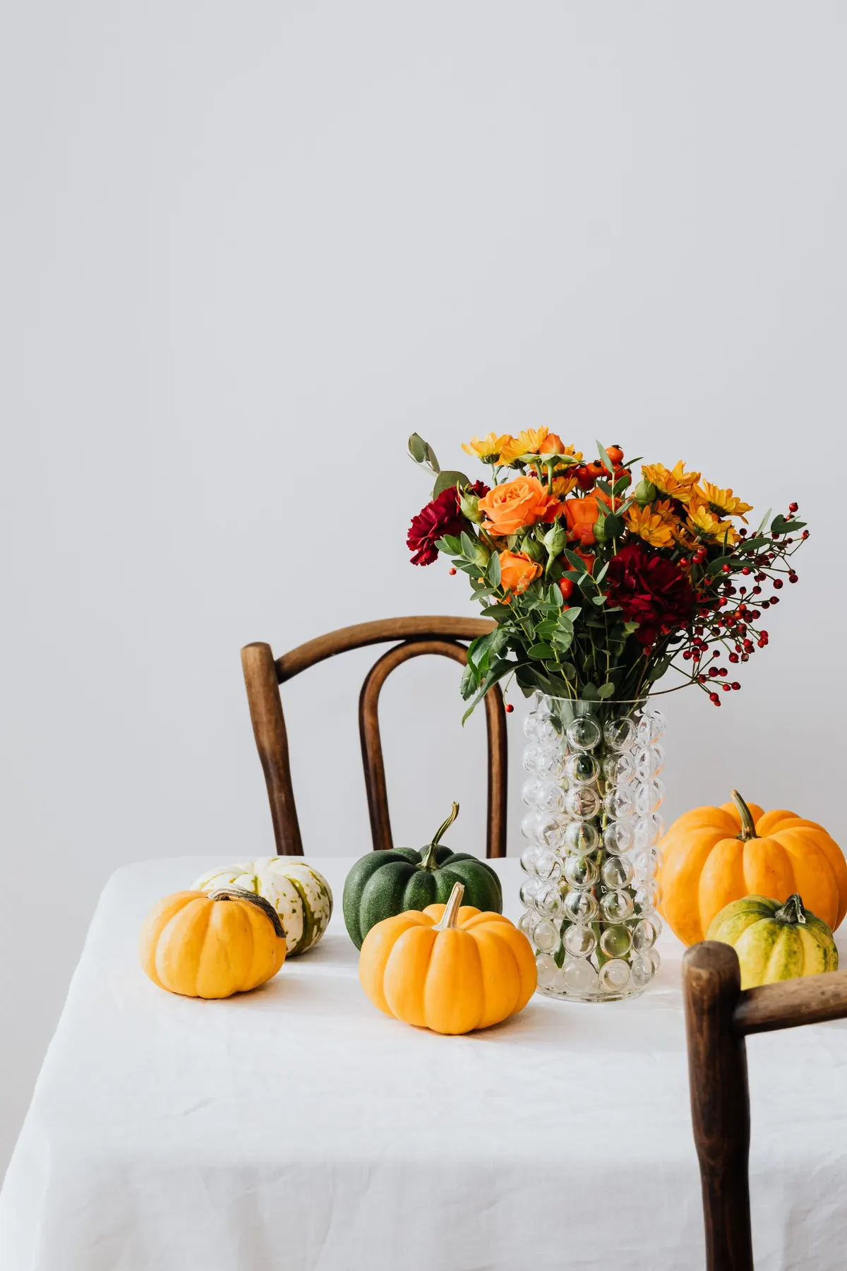 Small white-clothed table with a glass vase of orange roses and daisies, mini pumpkins and a bentwood chair
