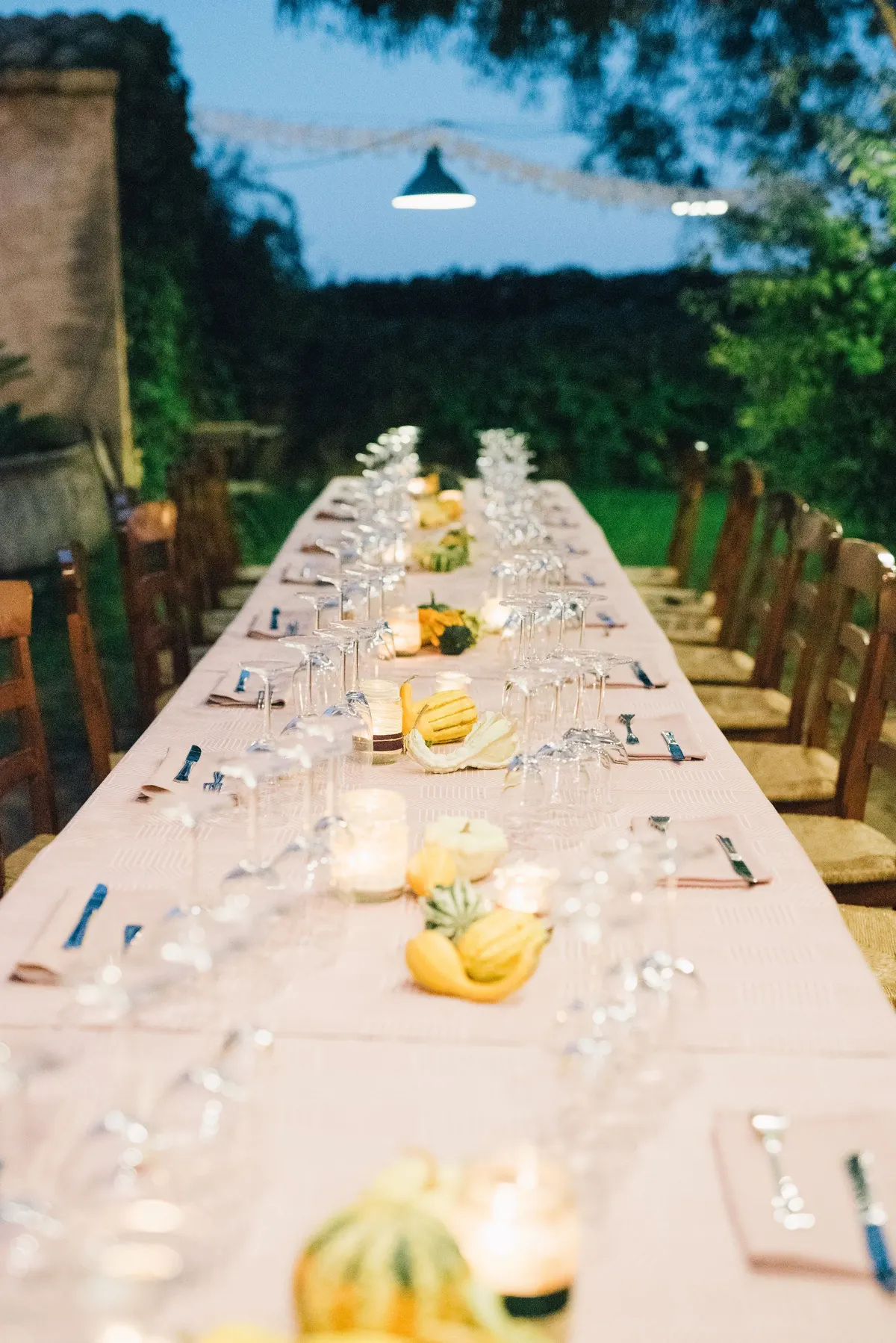 Long outdoor dinner table at dusk with pink linen, jar candles and low yellow and green gourds under string lights