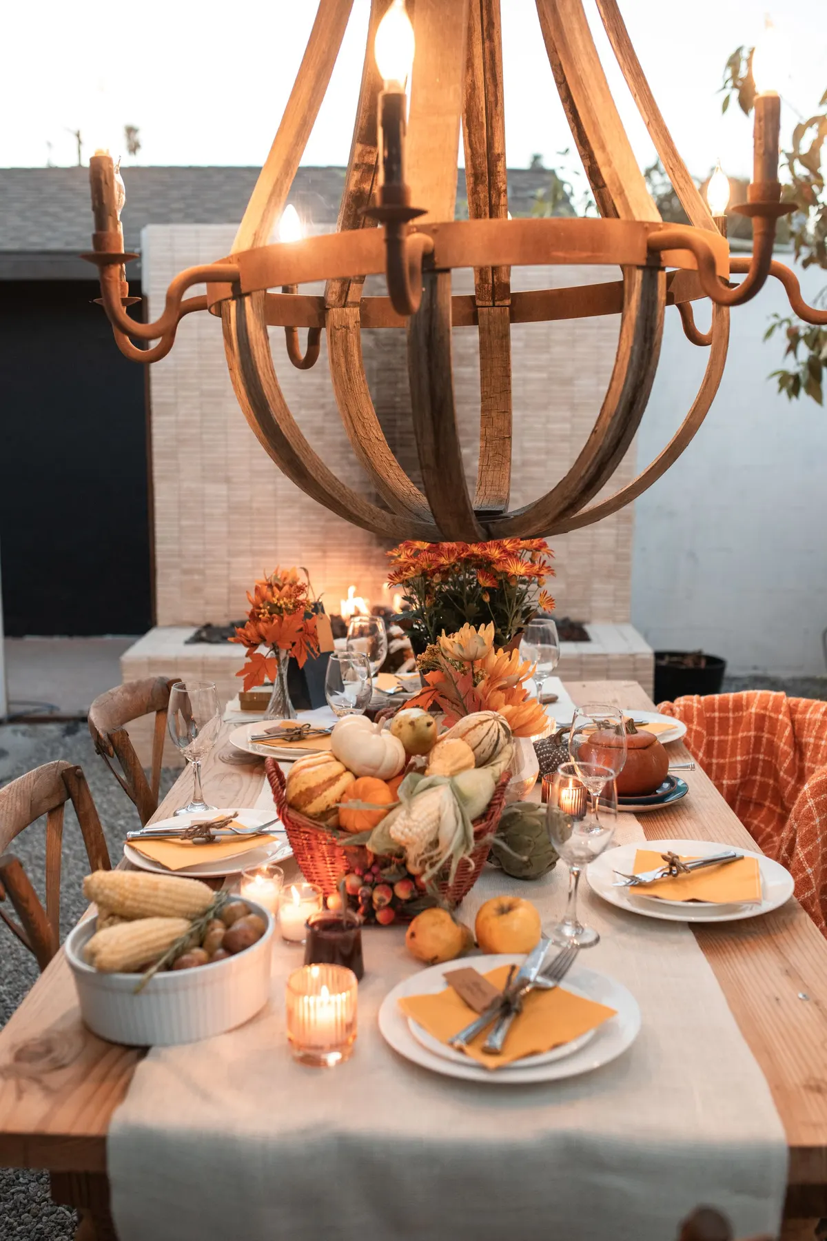 Outdoor wooden table under a barrel-stave chandelier with a gourd basket, mustard napkins, votives and orange flowers
