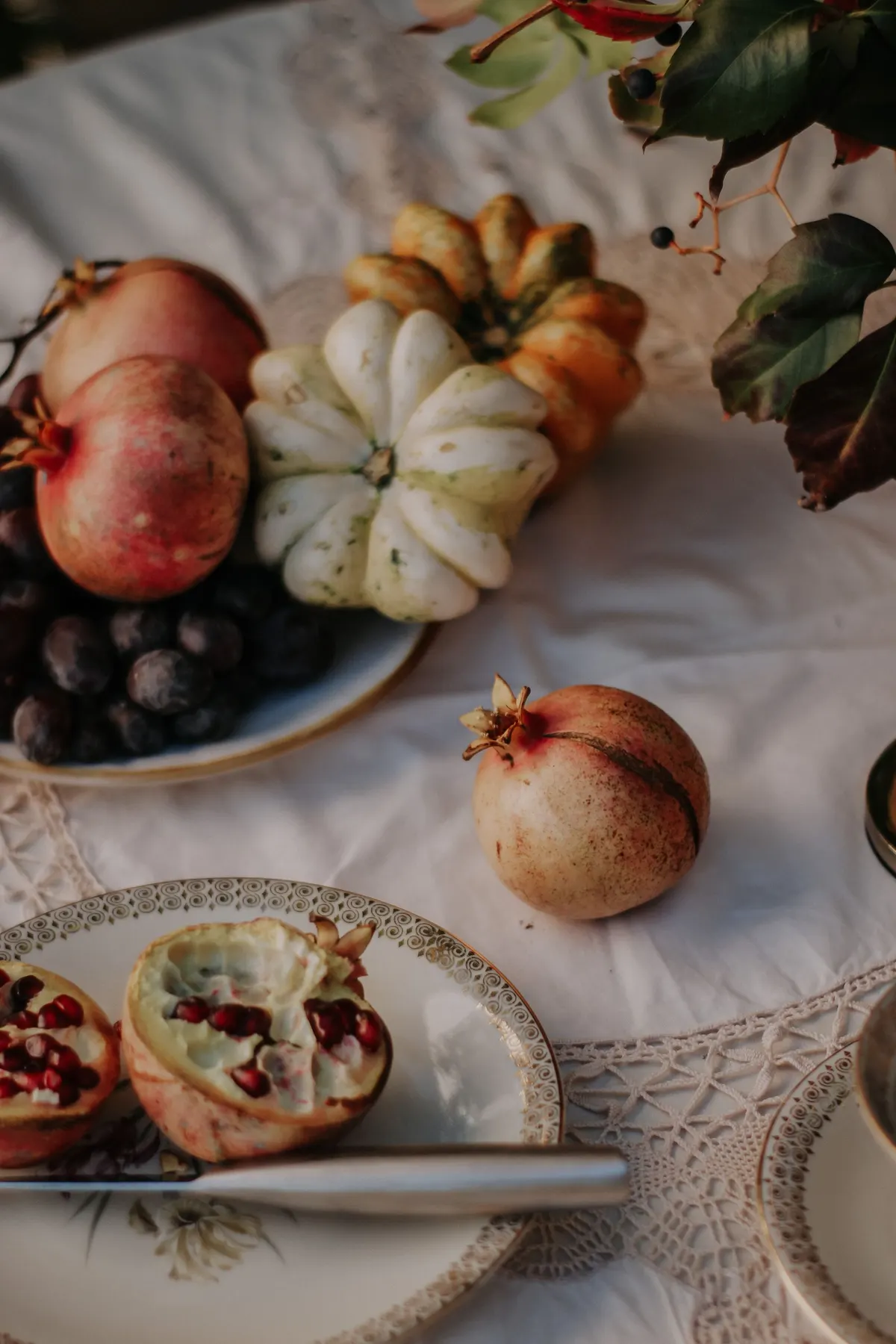 Pomegranates, plums and ribbed mini pumpkins on a gold-rimmed plate over a white lace tablecloth