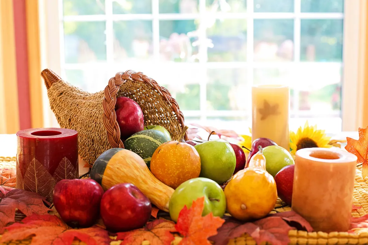 Wicker cornucopia spilling red and green apples and gourds beside three pillar candles in front of a window