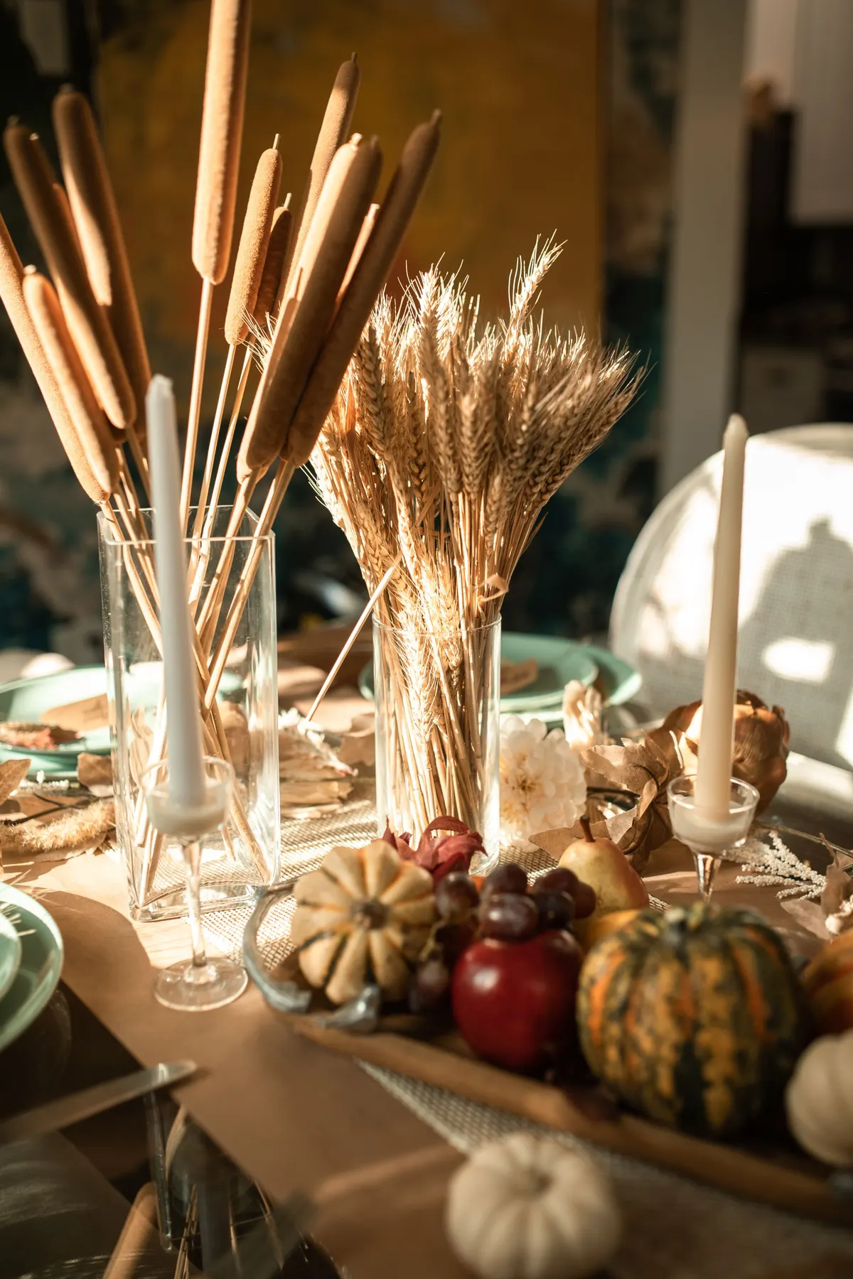 Tall glass vases of cattails and dried wheat beside white taper candles, mini pumpkins, apples and grapes on a paper runner