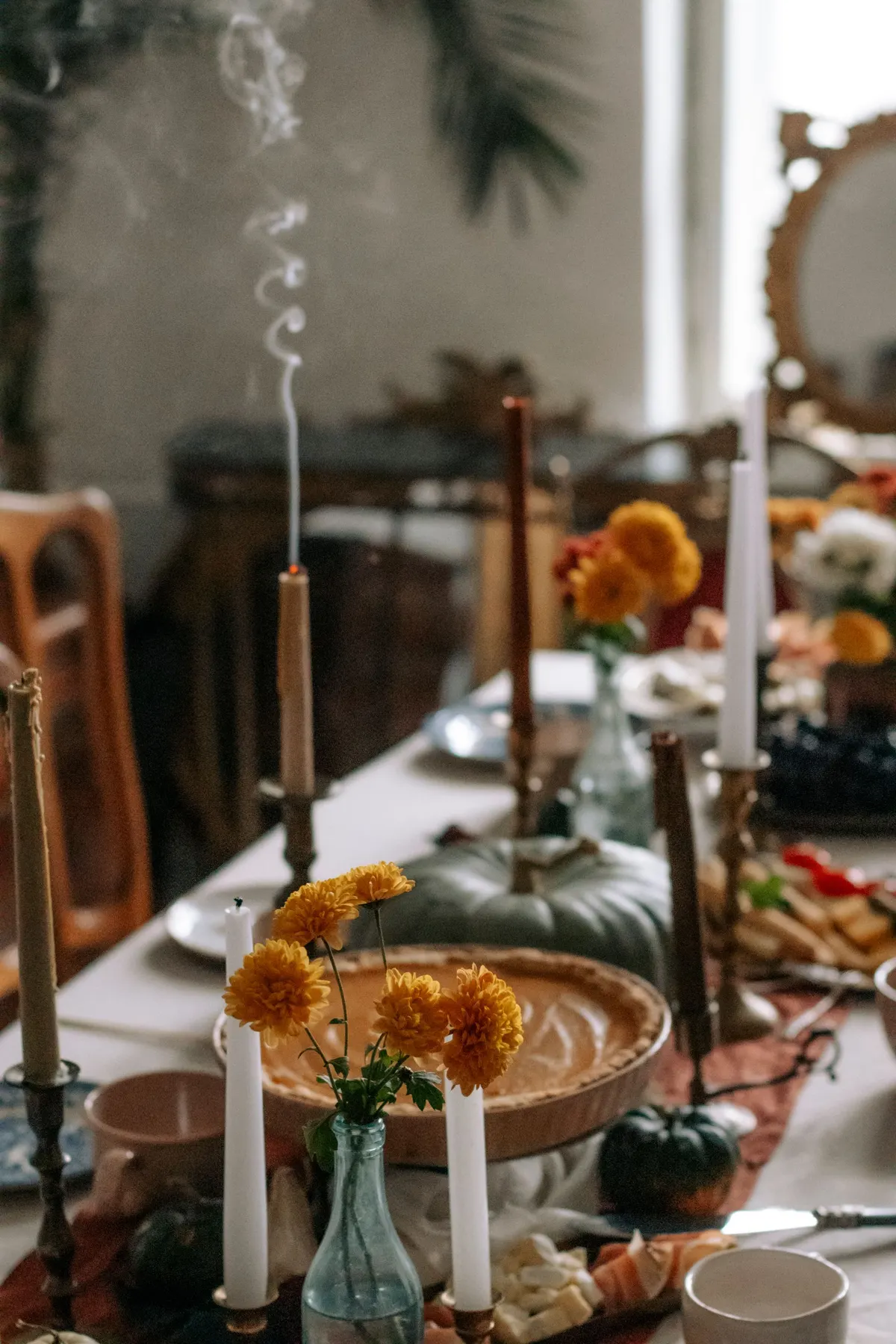 Long white-clothed table with taper candles in brass holders, mums in glass bottles, a pumpkin pie and small pumpkins