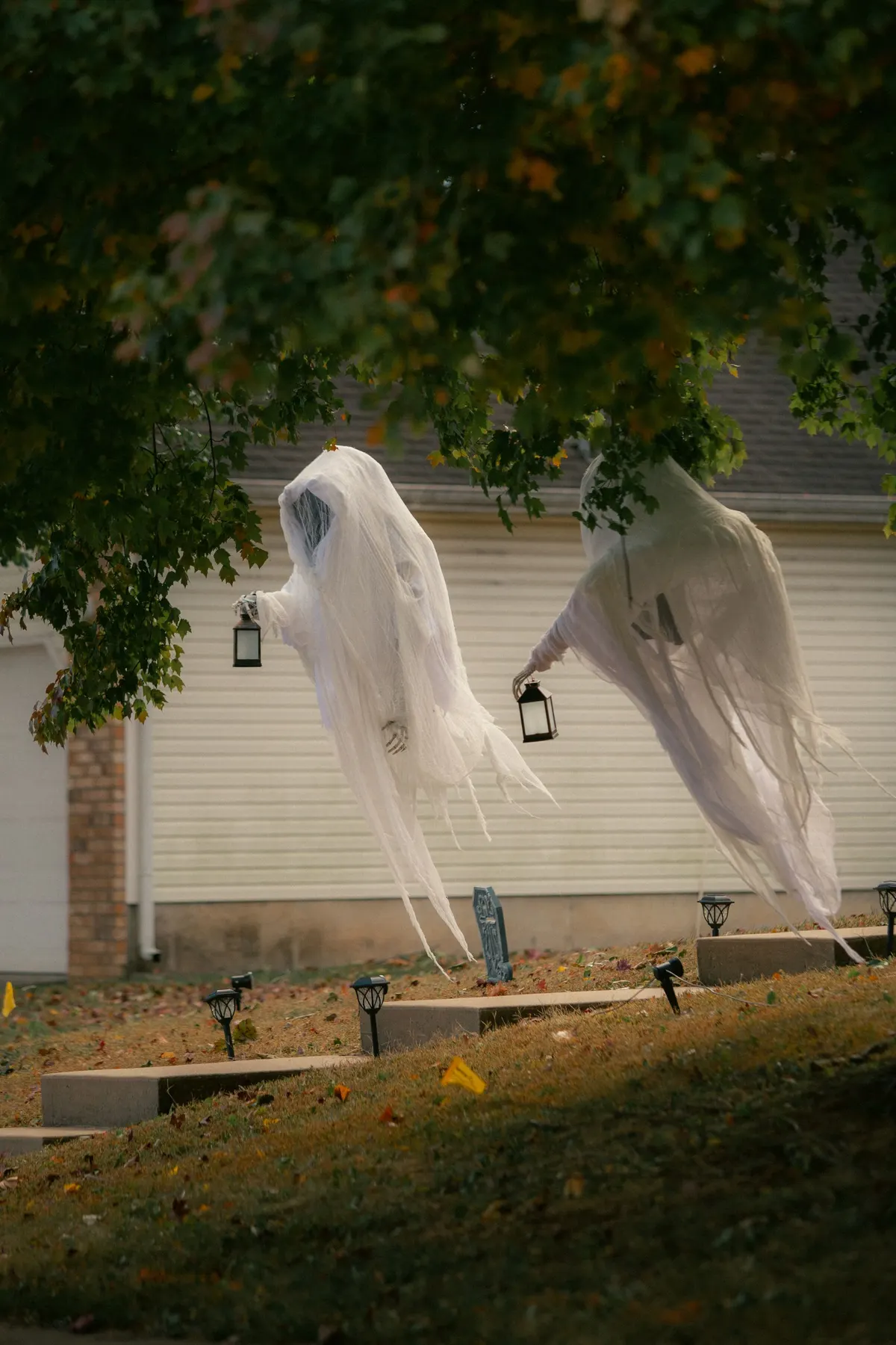 Two white gauze ghosts with frayed hems hanging from tree branches, each holding a small black lantern