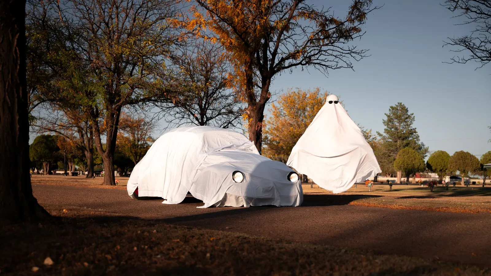 Classic car covered in a white sheet with round eye holes over the headlights, beside a tall cone-shaped sheet ghost