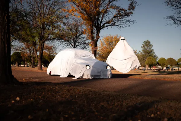 Classic car covered in a white sheet with round eye holes over the headlights, beside a tall cone-shaped sheet ghost
