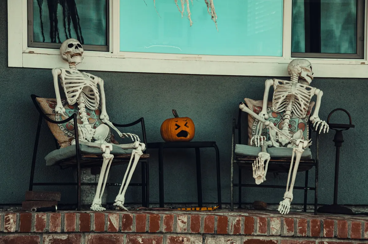 Two plastic skeletons in black iron chairs with pale green cushions chatting, with a small shocked-face jack-o'-lantern