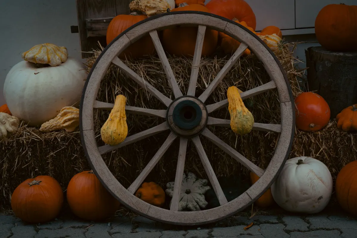 Wooden wagon wheel leaning on hay bales with yellow gourds in its spokes and orange and white pumpkins nearby