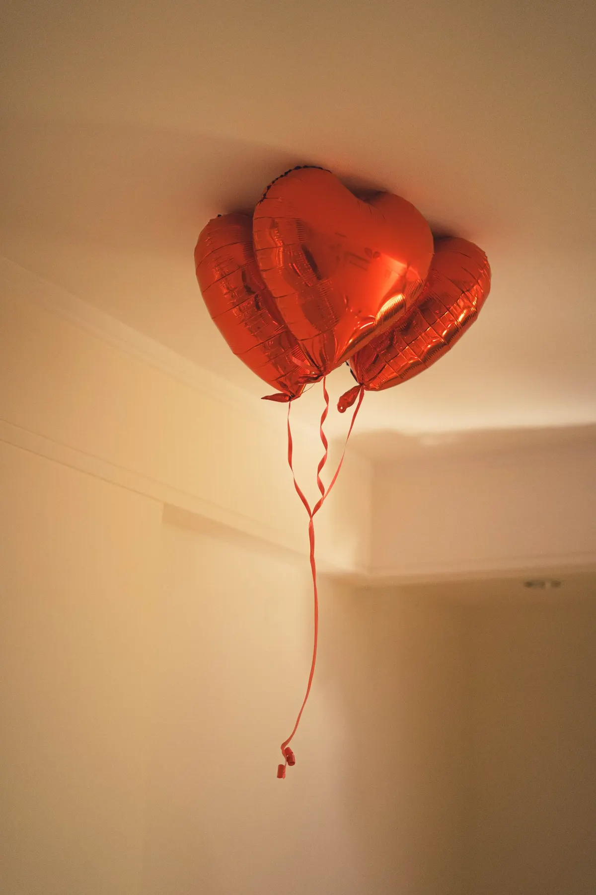 Three red heart-shaped foil balloons with red ribbons floating against a cream ceiling corner