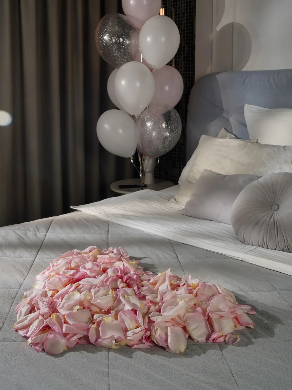 Bed with gray quilt and a large heart of pink and cream rose petals beside white and pink balloons