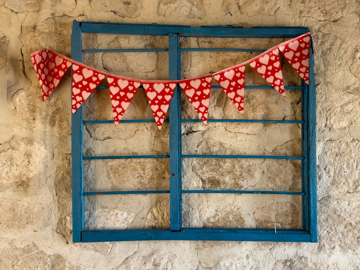 Eight red heart-print bunting flags hanging across a blue window frame on a rough beige stone wall