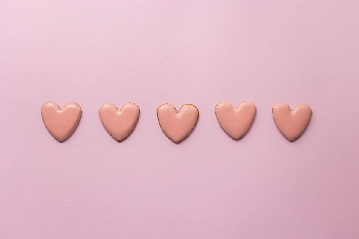 Five heart-shaped sugar cookies with glossy pale pink icing lined up in a row on a pink background