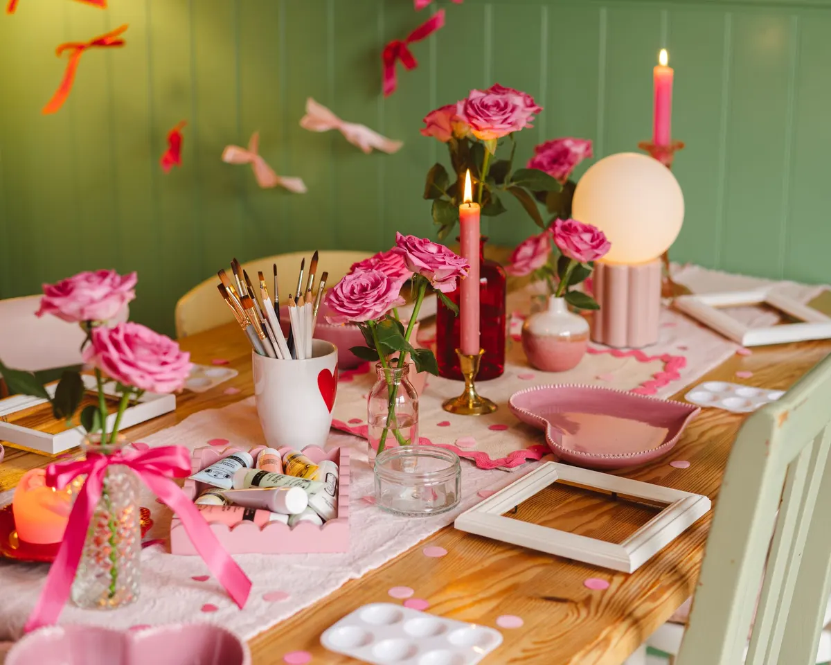 Craft table with pink roses, lit pink tapers, empty white frames, paint palettes, brushes in a mug and heart dishes