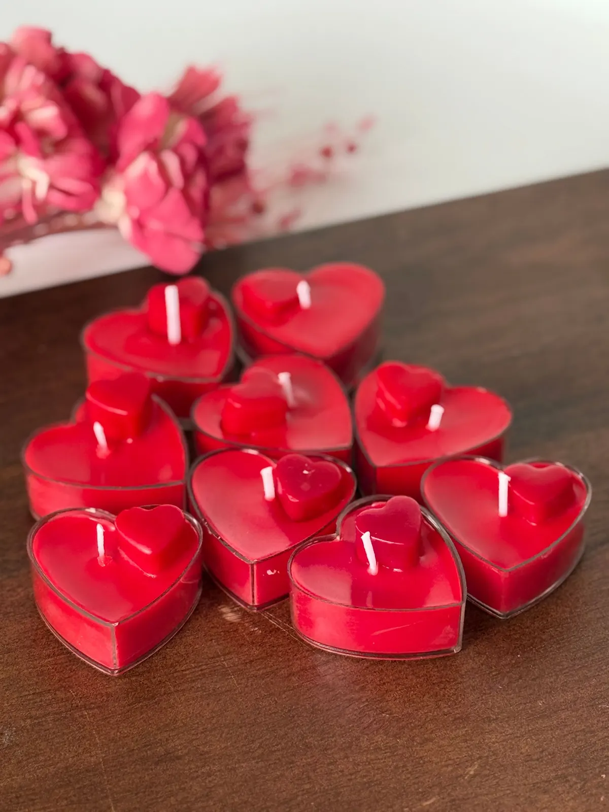 Eight red candles in clear heart-shaped holders on a dark wooden surface with blurred pink flowers behind
