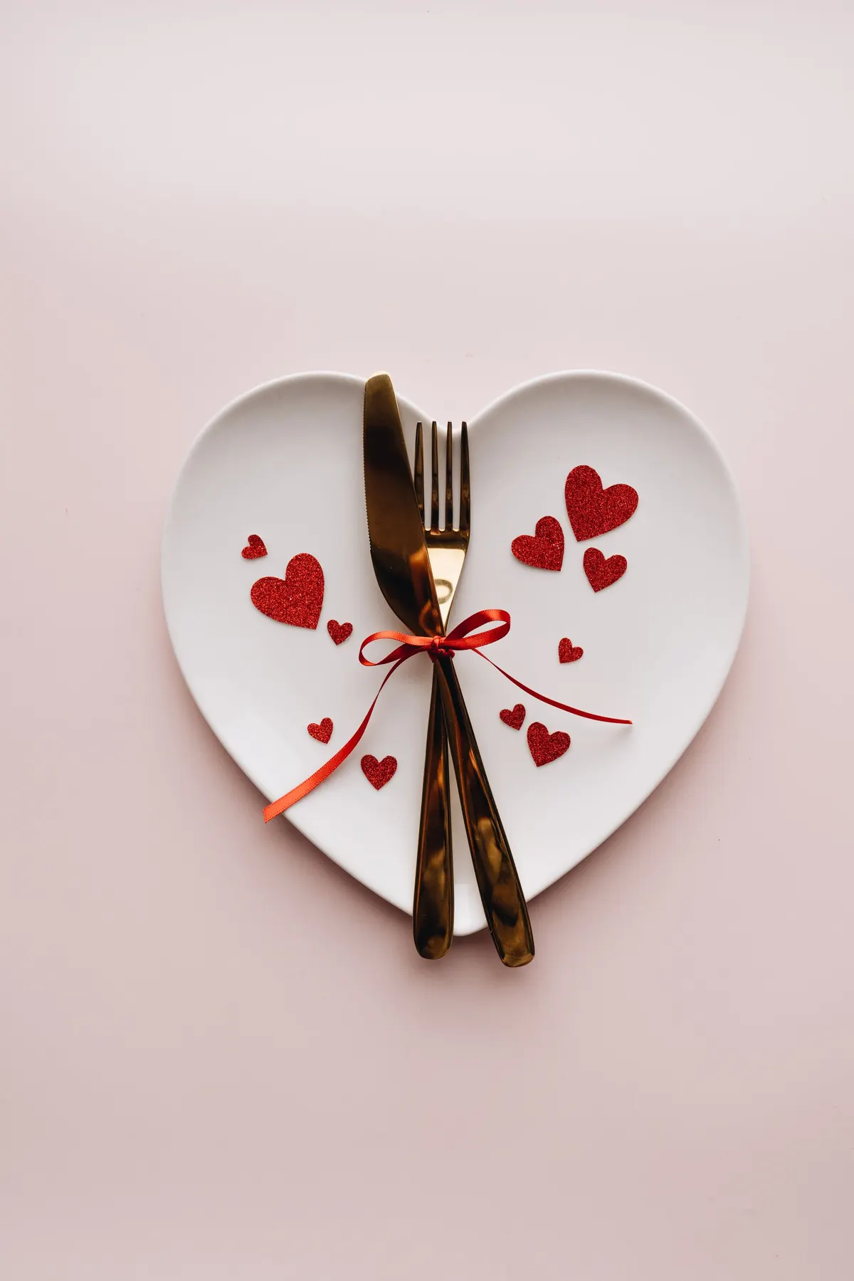 White heart-shaped plate with a gold fork and knife tied with red ribbon among red glitter hearts