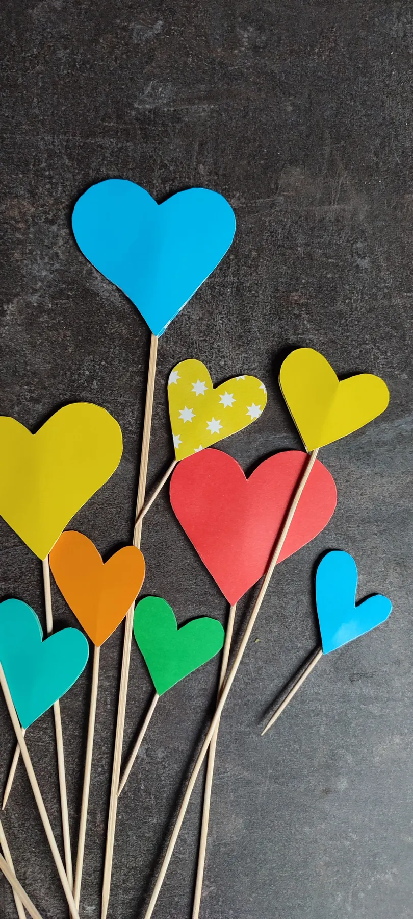 Bright paper hearts on thin wooden sticks in blue, yellow, red, orange, green and turquoise on dark gray