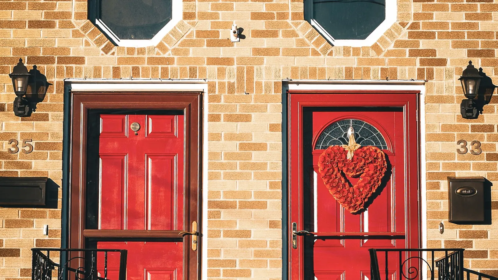 Brick house with two red doors, a red heart-shaped wreath on the right door and two black wall lanterns