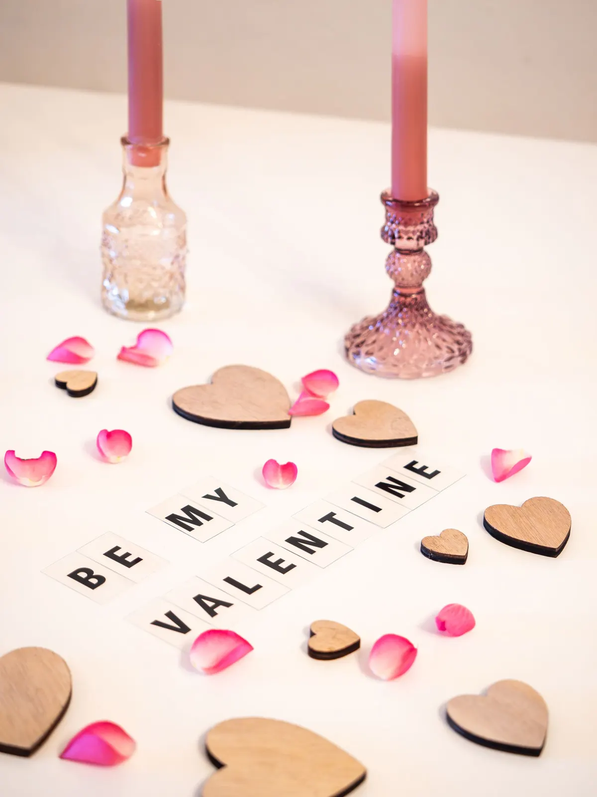 Letter tiles spelling Be My Valentine with two pink tapers, wooden hearts and pink and white rose petals