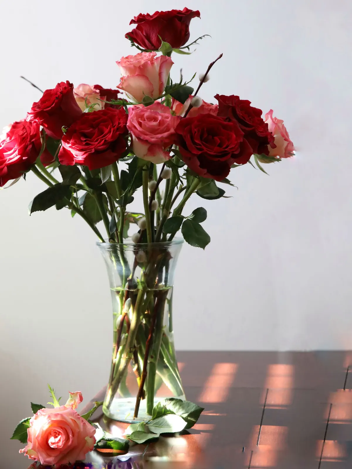 Clear glass vase of red and pink roses with fuzzy-budded twigs and one pink rose lying on a table
