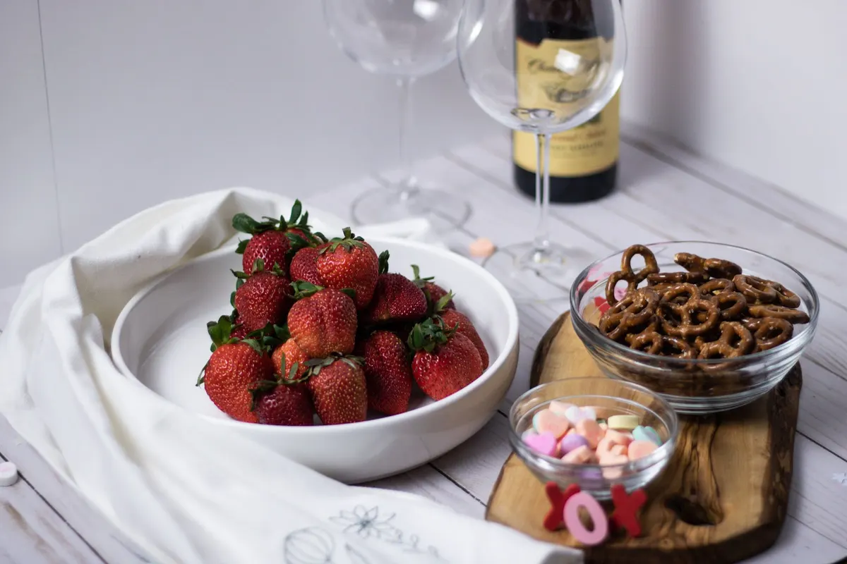 Wooden board with a white bowl of strawberries, a bowl of pretzels, pastel heart candies and two wine glasses
